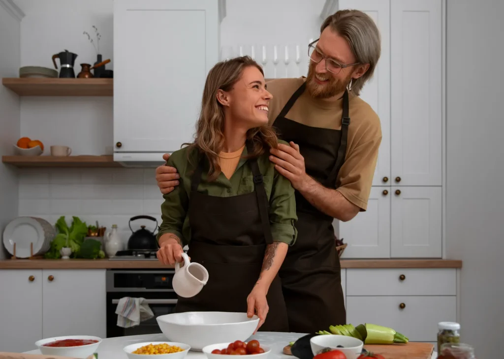 A woman smiles up at a man standing behind her in a modern kitchen. Both are wearing dark aprons. She’s holding a small white pitcher above a mixing bowl while he gently places his hands on her shoulders, smiling back at her. Fresh vegetables and ingredients are on the table in front of them.