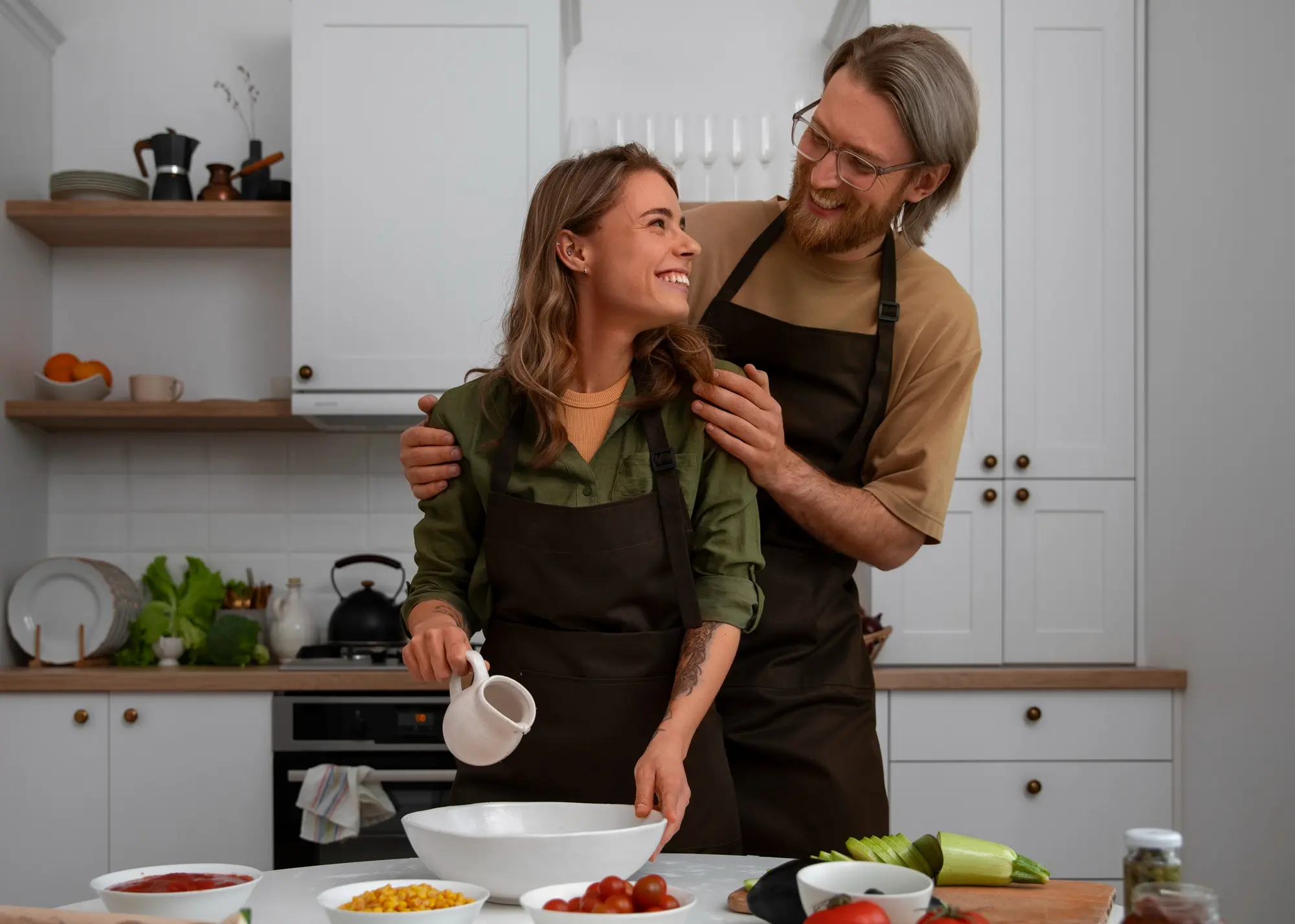 A woman smiles up at a man standing behind her in a modern kitchen. Both are wearing dark aprons. She’s holding a small white pitcher above a mixing bowl while he gently places his hands on her shoulders, smiling back at her. Fresh vegetables and ingredients are on the table in front of them.