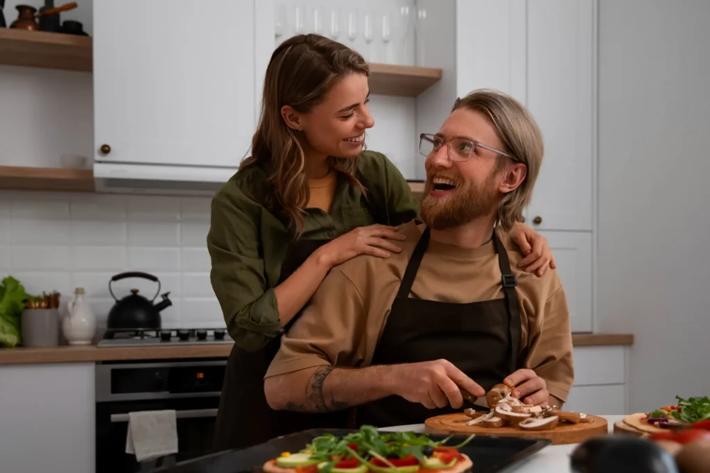 A woman stands behind a seated man in a home kitchen, both wearing aprons. She’s smiling warmly with her hands on his shoulders while he laughs and slices mushrooms on a cutting board. Colorful flatbreads with fresh toppings are visible in the foreground.
